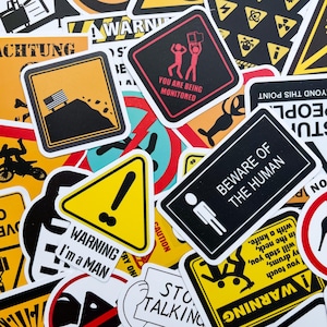Op de afbeelding: Een verzameling diverse rechthoekige en driehoekige stickers met gele, zwarte en rode waarschuwingsborden. De tekst bevat "YOU ARE BEING MONITORED", "BEWARE OF THE HUMAN", "WARNING I'm a MAN", "STOP TALKING" en "WARNING If you touch my drums, I will stab you in the neck, with a knife."