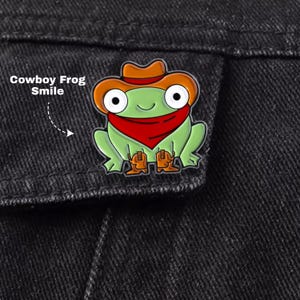 May include: A metal enamel pin featuring a light green frog wearing a brown cowboy hat, a red bandana, and brown cowboy boots. The frog has large white eyes with black pupils and a small smile. The text Cowboy Frog Smile is visible.