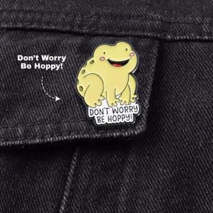 May include: A pale yellow enamel pin shaped like a smiling frog with small pink cheeks and dark spots on its back. The pin features the text "Don't Worry Be Hoppy!" in black lettering on a white background at the bottom of the frog figure.
