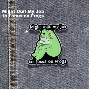May include: A green frog enamel pin featuring a cartoon frog sitting with its knees pulled to its chest. The pin includes black text that reads Might quit my job to focus on frogs, with the words positioned above and below the frog illustration.
