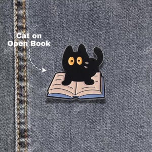 Könnte beinhalten: Eine schwarze Katzen-Emaille-Anstecknadel mit großen gelben Augen, die auf einem offenen Buch mit hellrosa Seiten und blauem Einband sitzt. Der Text Cat on Open Book steht in Weiß neben der Anstecknadel auf einem dunkelblauen Jeansstoff.
