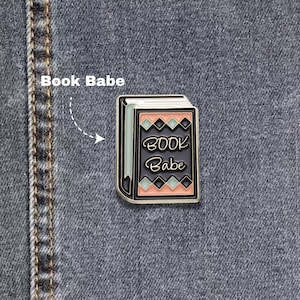 May include: A rectangular enamel pin shaped like a book with a black cover, light green pages, and peach-colored accents. The front cover features the text "BOOK Babe" in a white script font, surrounded by a geometric diamond pattern in black and green.