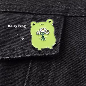May include: A lime green enamel pin shaped like a frog holding a bouquet of white daisies with yellow centres. The frog has a simple black line for a mouth and two small black dots for eyes. The text Daisy Frog is printed to the left of the pin.