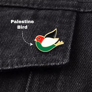 Peut inclure: Une broche en émail à bordure dorée en forme d'oiseau avec une tête rouge, un corps blanc, une partie inférieure verte et une aile noire. Le texte Palestine Bird est accompagné d'une flèche pointant vers l'oiseau sur un tissu en jean noir.