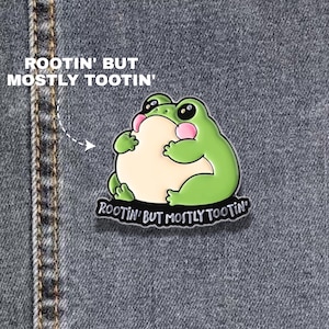 Könnte beinhalten: Ein grüner Emaille-Pin, der einen runden, sitzenden Frosch mit cremefarbenem Bauch und rosa Wangen zeigt. Der Pin enthält den Text "ROOTIN' BUT MOSTLY TOOTIN'" in weißer Schrift auf einem schwarzen Banner, platziert auf dunklem Jeansstoff.