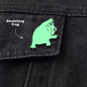 May include: A light green enamel pin shaped like a frog in a stretching pose, featuring black outlines and the text Stretching Frog printed in white next to it. The pin is attached to a black denim fabric surface with visible stitching details.