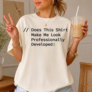 May include: An off-white short-sleeve t-shirt featuring the black printed text // Does This Shirt Make Me Look Professionally Developed followed by a small green bracket symbol. The shirt has a crew neckline and a relaxed fit.