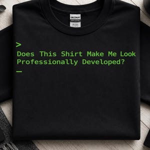 May include: A black crew neck t-shirt featuring a bright green graphic print that mimics a computer terminal interface. The text reads: > Does This Shirt Make Me Look Professionally Developed? - The shirt includes a Gildan brand neck label.
