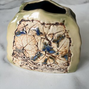 Könnte beinhalten: Eine handgefertigte Keramikvase mit rechteckiger Form und einzigartigem Blattdesign. Die Vase hat eine hellgrüne Außenseite mit einem detaillierten Relief von Blättern in Braun-, Creme- und Blautönen. Der Innenraum ist dunkel.