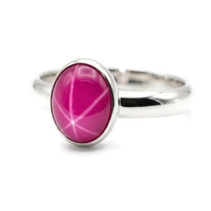 May include: A sterling silver ring featuring a large, oval-shaped, magenta-colored gemstone with a star-like pattern. The ring has a polished, smooth band and a bezel setting that securely holds the gemstone. The gemstone's color is a vibrant, deep pink.