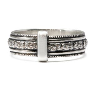 May include: A silver ring featuring a central smooth rectangular plaque flanked by bands of embossed floral patterns. The design includes beaded edges along the top and bottom, with an oxidized finish that highlights the intricate flower details.