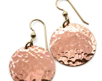 May include: A pair of copper-toned earrings featuring circular discs with a hammered, textured finish. Each disc is suspended from a simple metal ear wire hook, creating a dangling design that reflects light across the uneven, metallic surface.