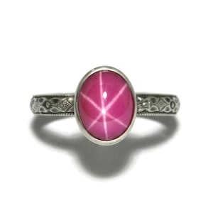 May include: A silver ring with an oval, star ruby gemstone. The pink ruby has a six-rayed star pattern. The ring band has an engraved design. The ring is set against a white background. The ring's design is ornate.
