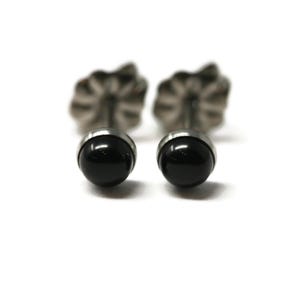 May include: A pair of stud earrings featuring smooth, round, polished black stones set in a metallic base. Each earring includes a scalloped, flower-shaped metal backing, providing a secure fit for these simple, circular, dark-colored jewelry pieces.