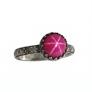 May include: A silver ring featuring a round, cabochon-cut, hot pink star gemstone set in a decorative, scalloped crown-style bezel. The band of the ring displays an intricate, embossed floral or leaf-like pattern along its entire outer surface.