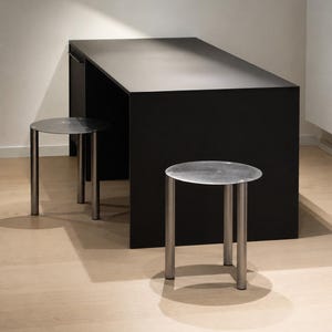 May include: A matte black rectangular table paired with two round stools featuring metallic silver circular tops and three cylindrical legs. The furniture set is arranged with one stool positioned in front of the table and another to the side.