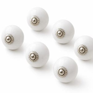 Puede incluir: Un juego de seis pomos esféricos blancos, cada uno con un acabado liso y brillante y una placa circular central de metal plateado con un patrón estriado en forma de rayos que rodea un pequeño domo central pulido de color plata.