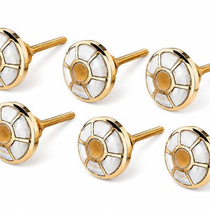 May include: A set of six round cabinet knobs featuring a polished gold-tone metal frame, a central amber-colored circular inlay, and a series of white, iridescent, petal-shaped segments arranged in a radial pattern around the center.