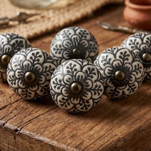 Op de afbeelding: Een set van zes ronde keramische kastknoppen met een zwart-wit bloemmotief. Elke knop heeft een glad, afgerond oppervlak met een centrale metalen schroefkop in antiek messingkleur en een ronde basis, gerangschikt in een groep.