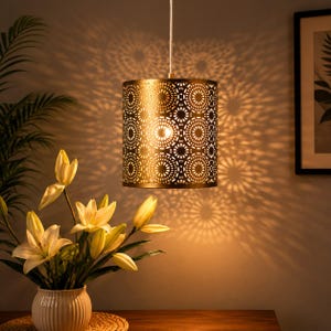May include: A cylindrical pendant lamp with a metallic gold finish and a repeating circular cut-out pattern that casts intricate shadows on the wall. The lamp hangs from a thin cord and illuminates a bouquet of white lilies in a ribbed vase.