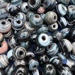 Könnte beinhalten: Eine Ansammlung runder Glasperlen mit glänzender Oberfläche und wirbelnden Mustern in Schwarz, Weiß, Grau und zartem Rosa. Jede Perle hat ein mittiges Loch und die Oberfläche weist eine glatte, reflektierende Textur in der gesamten Gruppe auf.