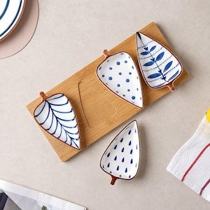 May include: A rectangular bamboo serving tray featuring three ceramic leaf-shaped dishes. Each dish has a white base with distinct blue patterns: one with dots, one with a central leaf vein, and one with a fern-like design. A fourth dish sits below.