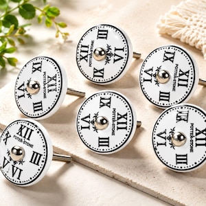 May include: A set of six round white ceramic cabinet knobs featuring black Roman numerals arranged like a clock face. Each knob has a central metallic silver-toned button and the text POTTER-STONE printed in black. A threaded metal bolt extends from the back.