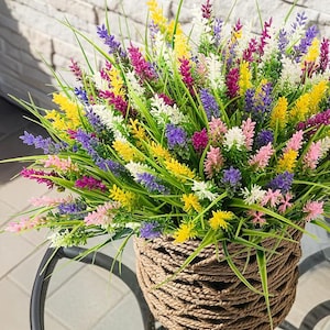 May include: A vibrant arrangement of artificial lavender flowers in a woven basket. The flowers are a mix of yellow, purple, pink, and white, with green grass-like stems. The basket is made of a light brown rope-like material.