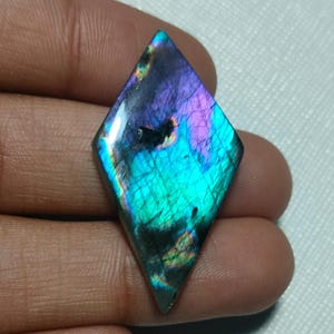 Op de afbeelding: Een ruitvormige labradoriet edelsteen met een gepolijst oppervlak met iriserende flitsen van levendig paars, elektrisch blauw en groenblauw, met natuurlijke interne breuken en een gladde, reflecterende afwerking.