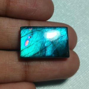 Pode incluir: Uma pedra preciosa de labradorite retangular com uma superfície polida e uma iridescência vibrante em tons de azul e verde-azulado, exibindo fraturas internas e um brilho distinto de luz rosa e laranja na sua estrutura cristalina escura.