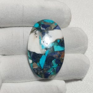 Könnte beinhalten: Ein ovaler Cabochon-Edelstein mit einem Mosaik-Inlay-Muster. Die Oberfläche zeigt eine Mischung aus polierten Fragmenten in den Farben leuchtendes Türkis, tiefes Marineblau, undurchsichtiges Weiß und Erdbraun mit glatter Oberfläche.