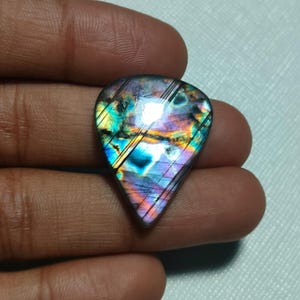 Puede incluir: Una piedra preciosa de labradorita en forma de lágrima con una superficie pulida que presenta destellos iridiscentes de azul, morado, amarillo y naranja. La piedra muestra distintas líneas diagonales grabadas en su cara, exhibiendo un juego de colores.