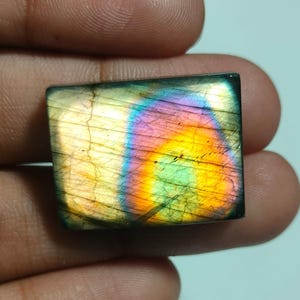 Puede incluir: Una piedra preciosa de labradorita rectangular con una superficie pulida que presenta destellos iridiscentes en amarillo, naranja, verde y morado. La piedra muestra fracturas internas naturales y un espectro de colores vibrante en su cara plana.