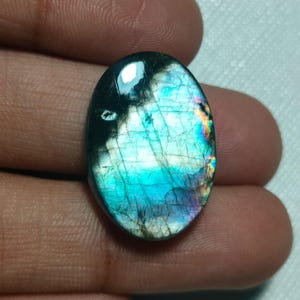 Könnte beinhalten: Ein ovaler Labradorit-Edelstein mit einer polierten Oberfläche, die irisierende Lichtreflexe in Blau, Grün und Gelb aufweist. Der Stein zeigt eine dunkle, halbtransparente Basis mit sichtbaren inneren Rissen und einem glatten, abgerundeten Rand.