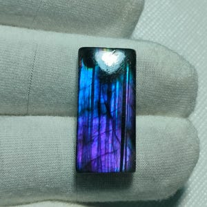 May include: A rectangular labradorite gemstone featuring a polished surface with vibrant iridescent flashes of deep blue and purple hues, displaying natural internal fractures and a smooth, flat finish across the entire stone face.