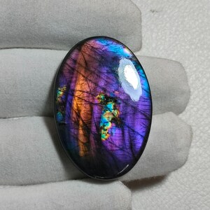 May include: An oval-shaped labradorite gemstone featuring a polished surface with deep purple, vibrant blue, and warm orange iridescent flashes. The stone displays a dark, semi-translucent base with fine internal fractures and a reflective sheen.