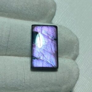 May include: A rectangular labradorite gemstone cabochon featuring a polished surface with a vibrant purple iridescence, visible internal fractures, and a distinct dark inclusion on the left side, set against a textured white fabric background.
