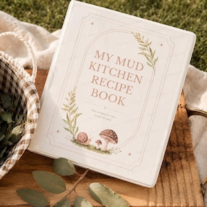 Peut inclure: Un livre de recettes crème intitulé "My Mud Kitchen Recipe Book" avec des illustrations botaniques de feuilles, d'un escargot et de champignons. Le livre est ouvert sur une surface en bois avec un panier et de la verdure en arrière-plan.