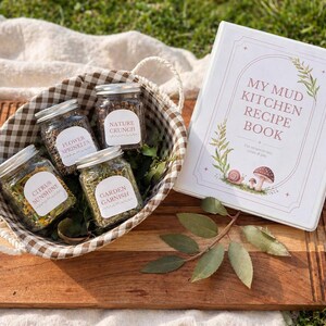 May include: A wooden serving tray displays a woven basket containing four glass jars of spices, labelled "Citrus Sunshine," "Flower Sprinkles," "Nature Crunch," and "Garden Garnish." A recipe book titled "My Mud Kitchen Recipe Book" is also present.
