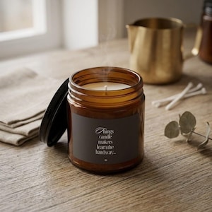 May include: A lit scented candle in a brown glass jar with a black lid. The candle has a white wick and a label with the text "Things candle makers learn the hardway...". A gold pitcher and cotton swabs are in the background.