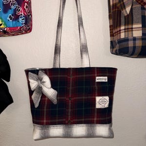 Puede incluir: Un bolso tote confeccionado con tela de franela a cuadros reciclada en tonos rojo, azul marino y verde. Presenta un panel inferior a cuadros blancos y negros, dos correas de hombro a juego, un lazo decorativo y dos etiquetas de marca visibles.