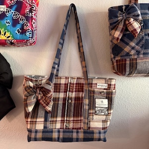 May include: A tote bag made from repurposed plaid fabric, featuring a large bow. The bag has a mix of red, white, and blue plaid patterns, with a pocket and a label that reads "BUCK CAMP". The handles are a contrasting blue and tan plaid.