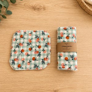 May include: Two square washcloths featuring a light green and white gingham pattern with a repeating peach fruit print. One cloth is laid flat, while the other is rolled and secured with a brown paper band labeled Handmade Washcloths.