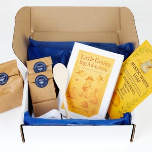 May include: A white box filled with baking ingredients. It includes brown paper bags marked "Flour", "Yeast", and "Salt", a wooden spoon, a book titled "Little Grain's Big Adventure", and a yellow packet of "Rustic White Loaf". Blue tissue paper lines the box.