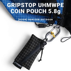 Pu&ograve; includere: Portamonete a scacchi bianchi e neri con cerniera nera e moschettone. La custodia &egrave; attaccata a un tessuto blu e nero. Il testo sulla custodia recita "HODR GEAR". Il testo "GRIPSTOP UHMWPE COIN POUCH 5.8g" &egrave; visibile.