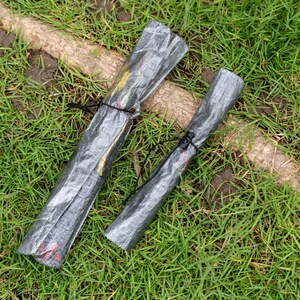May include: Two cylindrical items wrapped in translucent, crinkled gray plastic material. Each roll is secured with a thin black elastic cord tied around the center. The items are positioned diagonally, showing a textured, semi-transparent surface.