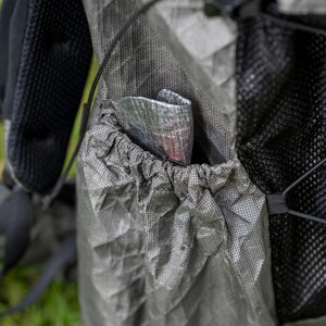May include: A gray, textured, ripstop fabric backpack side pocket with an elastic drawstring closure. A small, metallic, silver-toned, crinkled pouch is tucked inside the pocket. The backpack features black mesh panels and black straps.