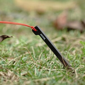 May include: A black metal tent stake with a pointed end and a flat top featuring a hole. A thin red cord is threaded through the hole at the top of the stake. The word HODR is printed in white capital letters on the side of the black tent stake.