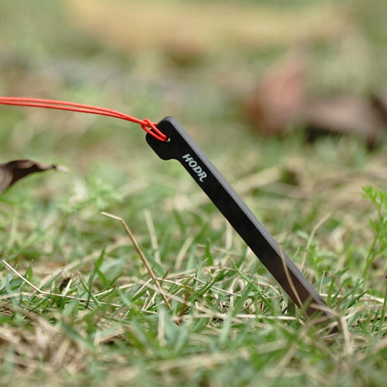 May include: A black metal tent stake with a pointed end and a flat top featuring a hole. A thin red cord is threaded through the hole at the top of the stake. The word HODR is printed in white capital letters on the side of the black tent stake.
