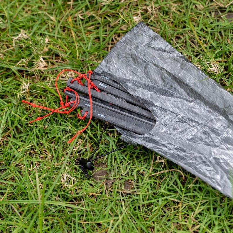 May include: A set of black carbon fiber tent stakes with red cordage, partially tucked inside a translucent gray crinkled storage pouch. The stakes feature a rectangular profile with the brand name HODR printed in white on the side of each unit.